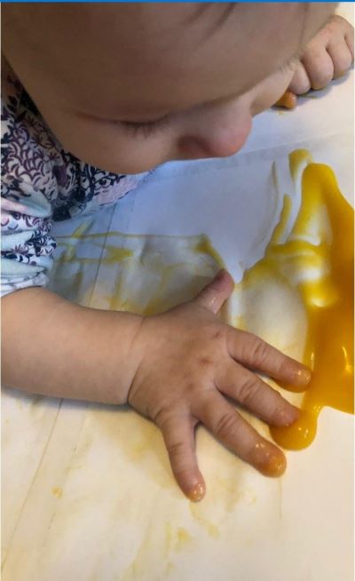 Pieni lapsi makaa lattialla vatsallaan ja maalaa kädellään valkoiselle kankaalle keltaisella soseella.